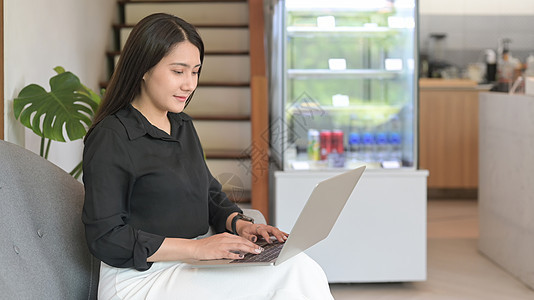 微笑的亚洲女性小企业家坐在她咖啡店的沙发上 使用笔记本电脑图片