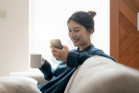 在家坐在沙发上时使用手机的年轻女性 充满欢欣鼓舞快乐房子黑发工具电话闲暇成人技术长椅微笑图片