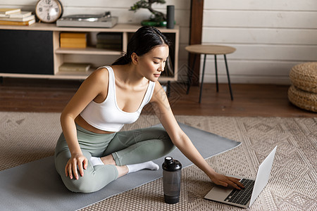 在家做瑜伽 在笔记本电脑上学习冥想课程 坐在地板垫子上坐着正姿势的年轻女性微笑哑铃房子女士技术运动成人房间身体有氧运动训练图片