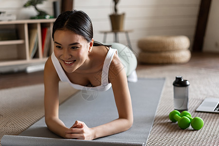 穿着运动服的运动型亚洲女孩在家锻炼 跟随视频运动指南 笔记本电脑上的在线健身教练 站在地垫上的木板上哑铃冥想技术女性房子训练互联图片