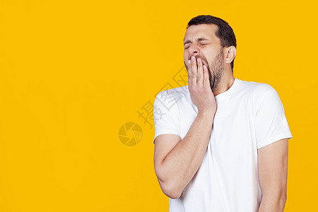 一个现代的年轻人 穿着白色T恤长胡子 在黄色背景下无聊无聊地打哈欠 厌倦了用手遮住嘴 无休止和沉睡图片