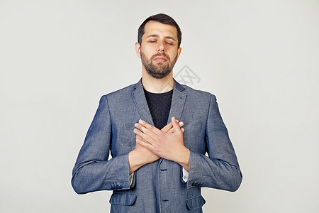 年轻的商务人士带着一个微笑的男人 留着胡子 穿着夹克 双手放在胸前 闭着眼睛 脸上挂着感激的手势 健康理念 灰色背景下的男人肖像图片