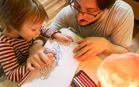 穿着灰色T恤的胡子男子和可爱男孩在家中共度时光时 用彩蜡笔绘画父亲男生童年爸爸微笑白色艺术桌子教育创造力图片