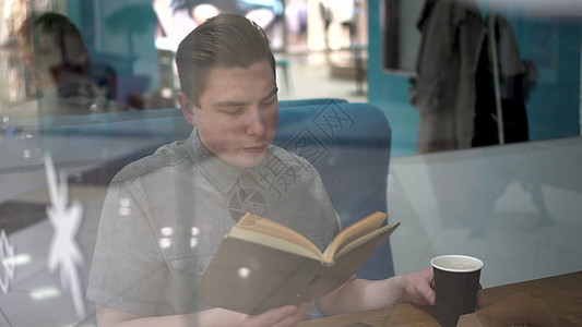 一名年轻人坐在咖啡馆的玻璃后面看一本书 他正在喝咖啡知识食堂学习男性大学写作咖啡青年流动餐厅图片