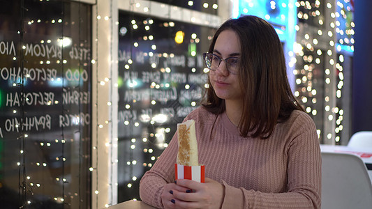 戴眼镜的年轻女子在一家餐馆吃卷饼 傍晚 一个女孩坐在咖啡馆里 窗边装饰着花环图片