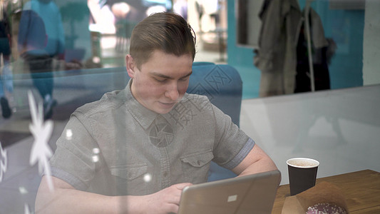坐在玻璃后面的年轻人拿着笔记本电脑坐在咖啡厅里 他喝咖啡和工作咖啡咖啡馆电子产品休息经理屏幕职业甜点专注沙发图片
