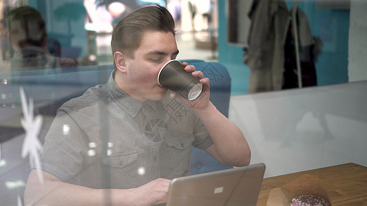 坐在玻璃后面的年轻人拿着笔记本电脑坐在咖啡厅里 他喝咖啡和工作商业午餐男性操作员远程自由职业者技术工具职业沙发图片