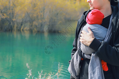 有吸引力的年轻父亲带着她的婴儿在户外吊带 男人背着她的孩子 在秋天的山湖中旅行 童装理念童年幸福公园儿子女孩女儿爸爸载体父母身份图片