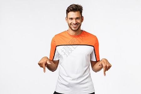 帅气 阳刚的西班牙裔运动员穿着运动 T 恤 穿着运动服 邀请您购买健身房会员资格 微笑神秘而高兴 向下指 介绍很棒的产品锻炼跑步图片