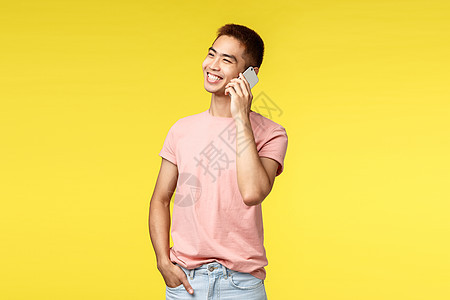 技术 通信和生活方式概念 身穿粉红色 T 恤 欢快的亚洲男性在手机上聊天 笑着 带着高兴的微笑看着别处 聊天 黄色背景的画像假期图片