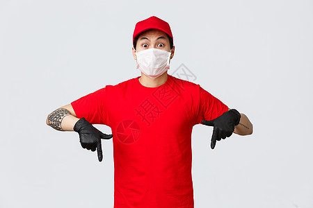 身穿红色制服帽和 T 恤的亚洲送货员惊奇而兴奋 用眼睛表达他的震惊和情绪 用防护手套和医用面具指着下方 展示新的促销优惠命令杂货图片