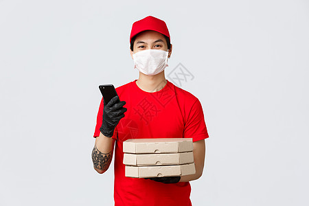 身着红色制服 拿着披萨盒和智能手机的友善亚洲送货员 送货服务建议在病毒爆发期间为安全在家的客户提供奖金 快递员戴医用口罩 手套肺图片