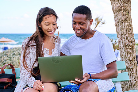 年轻夫妇看着笔记本电脑屏幕 坐在沙滩上的长椅上友谊幸福海滩男人女士公园男性假期微笑女孩图片