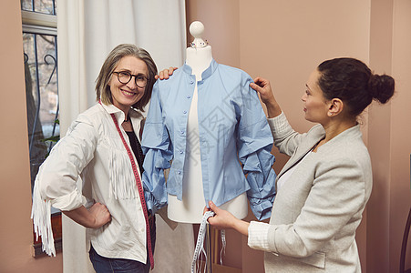 高级时装设计师微笑着与穿着新系列蓝色衬衫的假人合影 而她的同事裁缝则进行测量并致力于改进新服装的细节图片