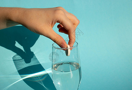 在蓝色背景下 一只手将一片溶解的冻状阿司匹林药片投进一杯水中剂量玻璃宿醉药品治疗帮助药物制药药理抗生素图片
