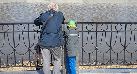 一个头发花白的祖父和他的孙子站在河堤上 靠在金属扶手上 在阳光明媚的春日里看着渔夫图片