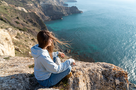 女性游客享受海山地貌的日落 坐在海面上的岩石上户外 穿着牛仔裤和蓝色连帽衫 健康生活方式 和谐与冥想 续瑜伽阳光天空自由旅行杯子图片