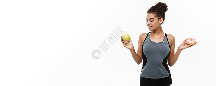 健康与饮食概念  美丽的运动性非裔美国人在甜甜圈和绿苹果之间做决定 孤立于白色背景损失食物肥胖甜点糖果水果女性营养身体重量图片