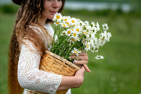 一位身穿白色连衣裙和棕色帽子的中年妇女手里拿着一个篮子 上面放着一大束雏菊 祝贺的野花季节甘菊植物群生活花束荒野洋甘菊花园乡村花图片