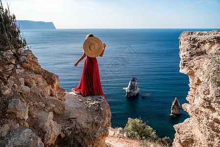 一个穿着飞红裙子的女人 在风中飘荡 和在海面背景下的草帽太阳旅行尾巴丝绸天空海滩幸福衣服成人女士图片