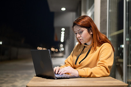 深夜 一位女士在夏季咖啡馆的笔记本电脑前远程工作 严肃的女孩坐在空荡荡的街道上的一张木桌旁学习 穿着运动衫的女性自由职业者图片