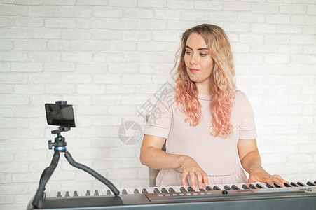 一位女士弹奏电子钢琴并在智能手机上创建视频博客 一个女孩用三脚架上的手机拍摄视频 女音乐老师远程工作课程教育歌曲乐器教程艺术家公图片