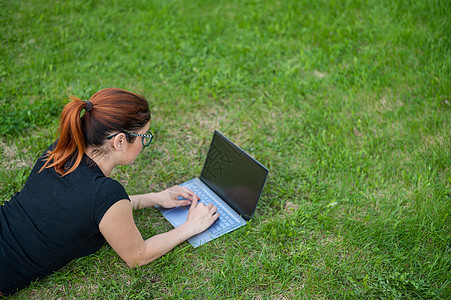 红头发的年轻女子在绿色草地上工作 自由女郎在户外笔记本电脑上印了指纹图片