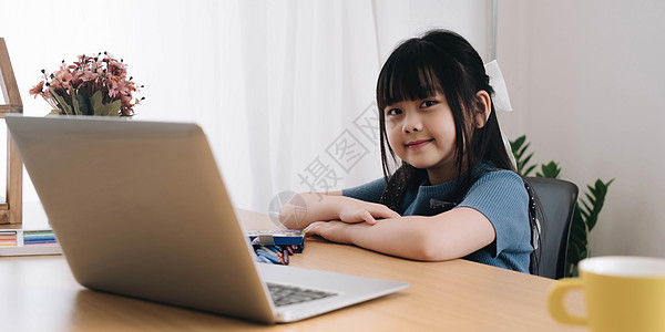 微笑的小女孩看着相机坐在桌前在家学习可爱的孩子做作业拿着笔做笔记看网络研讨会图片