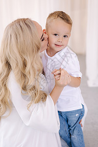 妈妈亲吻她的儿子 穿着优雅的白色礼服的女士在演播室里给摄影师摆姿势 白发背景 (笑声)图片
