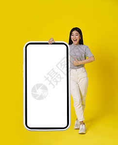 迷人的亚洲女孩竖起大拇指 站在巨大的智能手机旁 智能手机上有白色屏幕 穿着黄色背景的休闲装 很开心 自由空间模拟 植入式移动应用图片