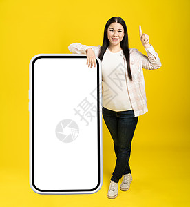 亚洲女性指着一根手指 注意力靠在巨大的 巨大的智能手机上 屏幕空白 穿着黄色背景的休闲装 自由空间模型图片