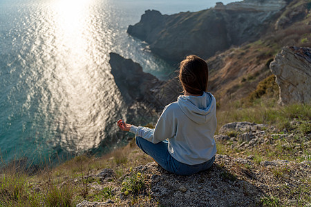 女性游客享受海山地貌的日落 坐在海面上的岩石上户外 穿着牛仔裤和蓝色连帽衫 健康生活方式 和谐与冥想 续太阳阳光杯子手势成人手印图片