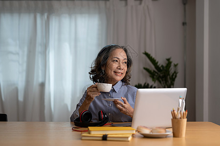 快乐快乐的成熟笔记本电脑用户亚洲女性在线聊天 打字 通过咖啡 茶进行视频通话 在沙发上放松 享受在家工作的资深自由职业女性图片