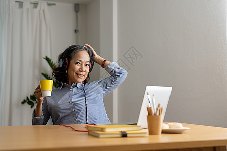 快乐的穿着耳机的高级亚洲女性 在舒适的家中客厅用笔记本电脑收听歌曲 看着相机图片