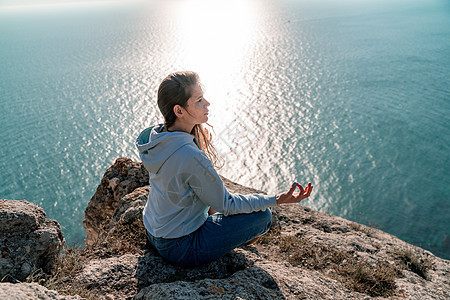 女性游客享受海山地貌的日落 坐在海面上的岩石上户外 穿着牛仔裤和蓝色连帽衫 健康生活方式 和谐与冥想 续天空喜悦沉思假期瑜珈自由图片