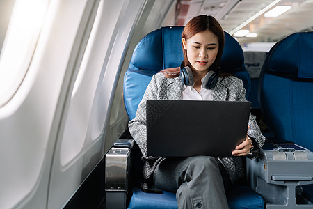 在飞行 旅行和商务概念期间 亚洲青年女子在飞机头等舱靠近窗户窗口的飞机上使用笔记本电脑戴耳机空气成人班级男人女孩工作运输航班女性背景