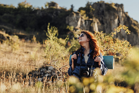 戴太阳镜 带数码相机和背包 带野营垫的年轻旅行者女性站在山上 女性徒步旅行者在她的单人旅行中享受自由图片