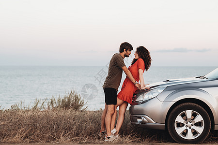 年轻情人和妇女共同站在日落海景海的背景的SUV汽车旁(SUV)图片