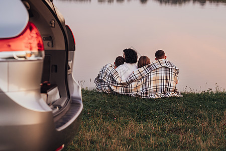 四名成员家庭坐在日落湖边的草原上 母亲和父亲与两个孩子一起享受周末大路之旅图片