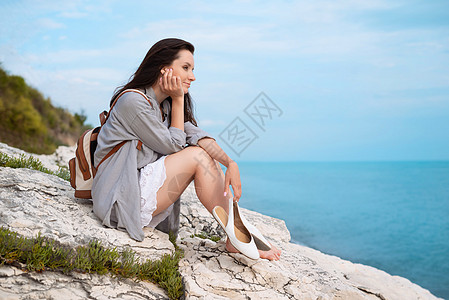 一个漂亮的旅游女孩 背着时髦的背包 穿着夏装 正坐在海边的一块白色岩石上自由蓝色女士海岸假期海滩冒充休息笑声旅行图片