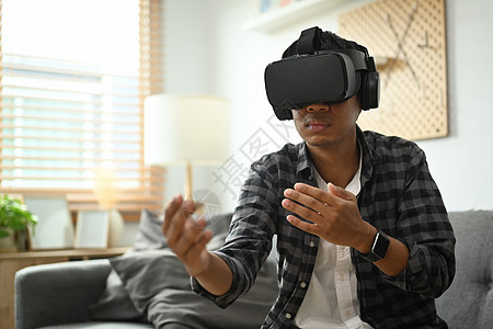 年轻的亚洲男子坐在沙发上 在VR玻璃耳机里玩电子游戏图片