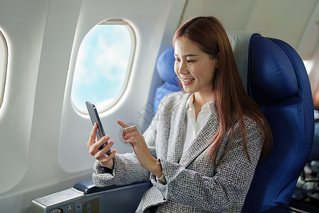 一个微笑着的亚洲女商务人士在飞机上使用她的手机的肖像行李企业家经理乘客游客手提箱旅行护照人士飞机场图片