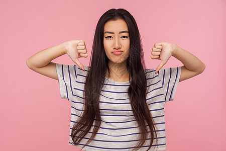 粉红色背景的亚洲年轻女性肖像女士批判悲伤鬼脸拇指条纹女孩愤怒长发厌恶图片