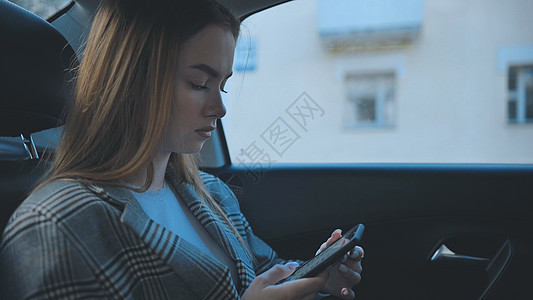 一个年轻女孩坐在汽车的后座上 看她的智能手机车辆奢华女士技术出租车电话轿车旅行药片乘客图片