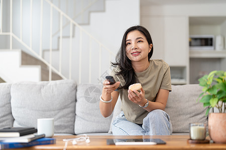 一位美丽的亚洲女性在家中坐在沙发上 用遥控器搜索频道观看电视的图像 笑声展示电影女士房间娱乐程序乐趣闲暇女孩技术图片
