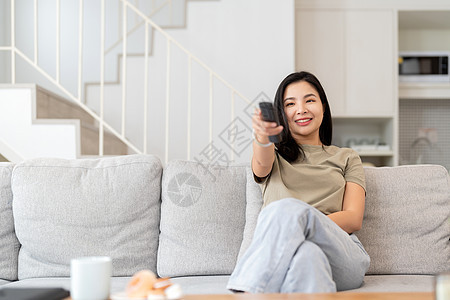 一位美丽的亚洲女性在家中坐在沙发上 用遥控器搜索频道观看电视的图像 笑声女士电影技术娱乐控制男人房间手表夫妻房子图片