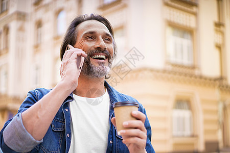 快乐旅行的成熟大胡子男人用手机说话 用一次性纸杯喝咖啡 站在户外穿着牛仔裤衬衫的旧城市背景中 自由职业者旅行人图片