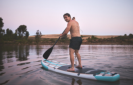 年轻活跃的白人男子微笑着 在大自然的湖上用他的站立桨板 年轻男性在黎明时分在桨板上享受夏日 当你在水中划桨时 生活会更美好图片