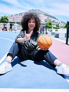 时髦的西班牙裔年轻女性用手和手指做出摇滚手势 在球场上拿着篮球 有爆炸头的快乐的年轻女人坐在篮球场上展示手势图片