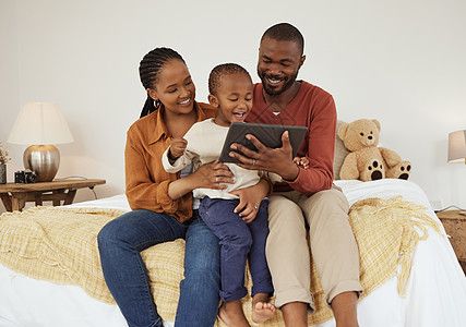 可爱的非洲裔美国小男孩和父母坐在床上使用数字平板电脑 年轻的父母看着兴奋的蹒跚学步的儿子玩游戏或在线看卡通电影图片